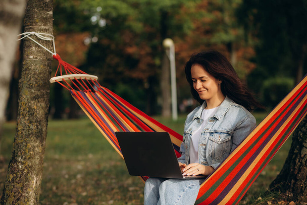 Junge schöne Frau liegt in einer Hängematte mit Laptop in einem Garten. Grüner Hintergrund. Bäume. Telearbeit. Fernarbeit. Hochwertiges Foto - Foto, Bild