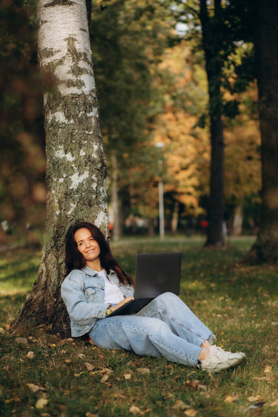 Γυναίκα που κάθεται κάτω από το δέντρο στο πάρκο και δουλεύει στο λάπτοπ και πίνει καφέ. Υψηλής ποιότητας φωτογραφία - Φωτογραφία, εικόνα