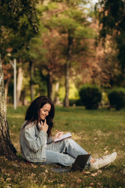 Hermosa estudiante guapa sentada bajo el árbol en el parque escribiendo en el ordenador portátil. Educación y concepto de trabajo remoto, espacio de copia. Foto de alta calidad - Foto, imagen