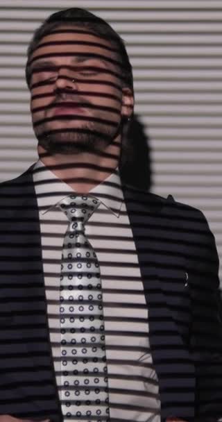 cooler Modeunternehmer, der zur Seite schaut, während er einen marineblauen Anzug zuknöpft, verführerisch die Lippen berührt und vor Jalousien auf grauem Hintergrund posiert - Filmmaterial, Video