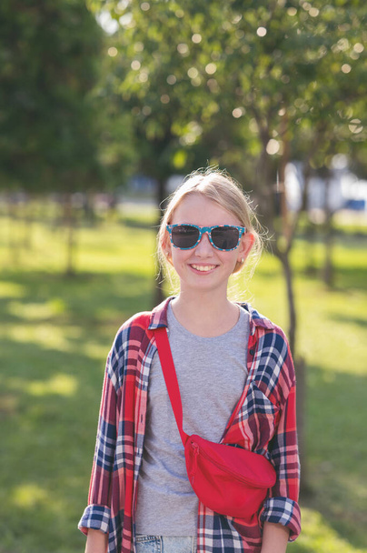 Retrato de una hermosa adolescente con una camisa a cuadros y gafas de sol en un parque de la ciudad sobre un fondo de árboles en un día soleado de verano. - Foto, imagen