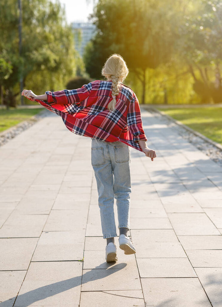 Дівчина в плетеній сорочці стрибає і біжить по стежці в міському парку в сонячний літній день. Концепція підліткового свята. - Фото, зображення