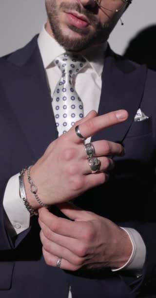 крупним планом відео крутого елегантного чоловіка в окулярах, що регулює браслети і костюм, бути крутим, дивитися в бік і класти руки в кишені на сірому фоні - Кадри, відео
