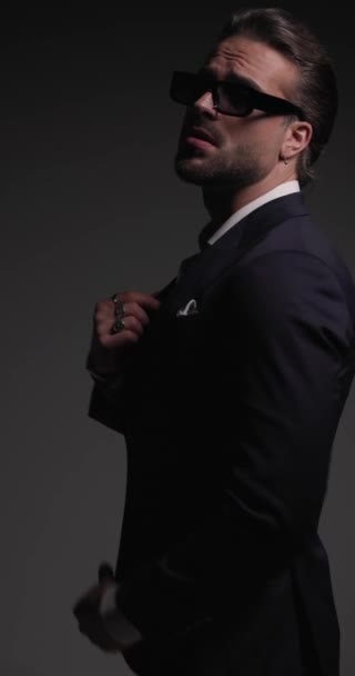 vista lateral video de sexy barbudo hombre de negocios con gafas de sol en movimiento, frotando las palmas, ajustando el traje, tocando el hombro y mirando a un lado en las luces de flash fondo gris - Metraje, vídeo