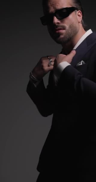 cool fashion man with sunglasses adjusting suit and confidently posing in front of flash lights on grey background - Footage, Video