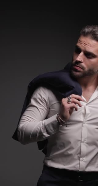 вертикальное видео сексуального бизнесмена касающегося подбородка и задумчивого, держащего пиджак через плечо и смотрящего в сторону на сером фоне - Кадры, видео