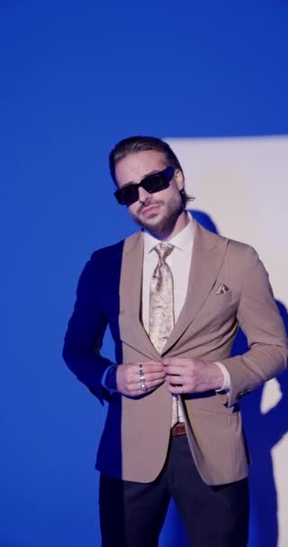 элегантный бизнесмен высокого класса в солнечных очках застегивая коричневый костюм, держа руку в кармане и глядя в сторону на сером фоне - Кадры, видео