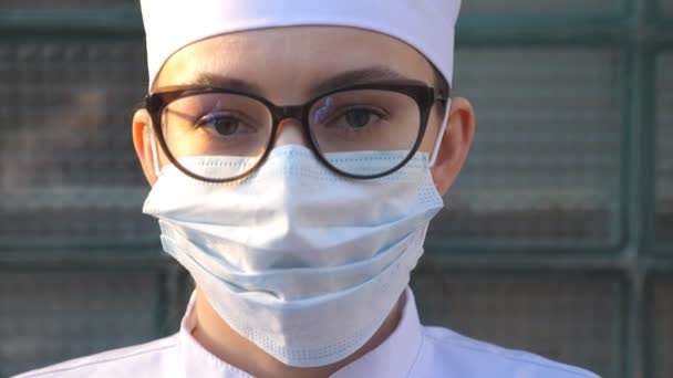 Retrato de jovem médica com máscara facial médica olha para a câmera. Trabalhador médico usando máscara protetora contra vírus. Conceito de saúde e segurança da pandemia de coronavírus. COVID-19
. - Filmagem, Vídeo