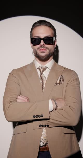 vídeo vertical de caballero elegante guapo con gafas de sol cruzando brazos y mirando a un lado delante de proyector redondo sobre fondo gris - Metraje, vídeo