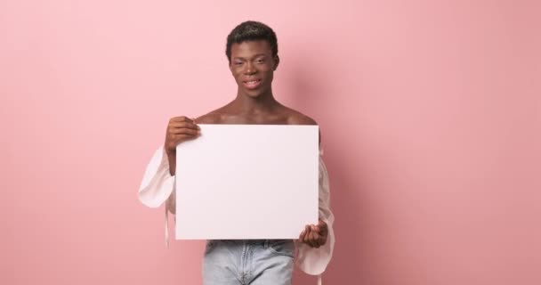 Persona transgender africana sorridente mentre tiene un poster bianco vuoto su uno sfondo isolato. Concetto pubblicitario. - Filmati, video