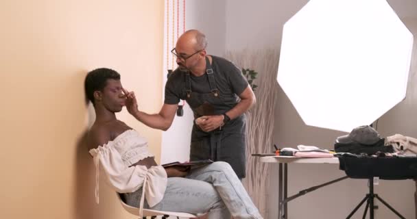 Full length of male makeup artist applying lipstick on lips of African American drag queen wearing pink top while sitting on chair in studio against beige backdrop - Footage, Video