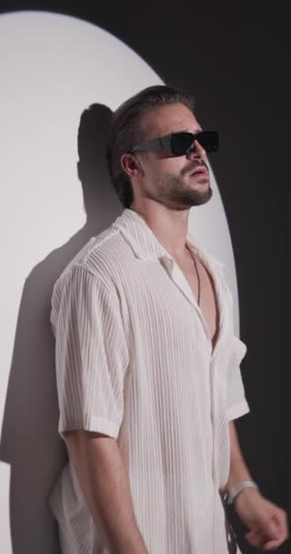 hűvös fiatal férfi fehér ing néz előre, és állítsa napszemüveg, dörzsölés tenyér és magabiztos szürke háttér - Felvétel, videó