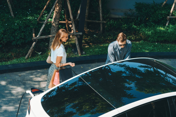 Young couple travel with EV electric car charging in green sustainable city outdoor garden in summer shows urban sustainability lifestyle by green clean rechargeable energy of electric vehicle innards - Photo, Image