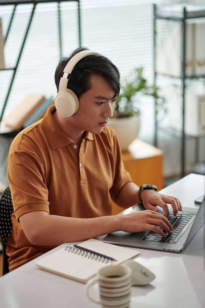 Νέοι βιετναμέζοι δοκιμαστής ακούγοντας μουσική όταν εργάζονται στο γραφείο στο γραφείο στο σπίτι - Φωτογραφία, εικόνα