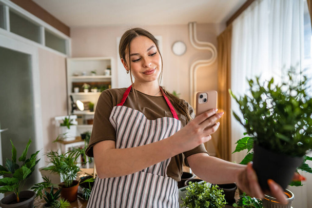 Одна женщина молодого поколения Z взрослых кавказских женщин заботиться о своих растений дома фотографируя цветочный горшок с помощью смартфона мобильного телефона отправить фотографии в социальных сетях или в качестве сообщения реального человека - Фото, изображение