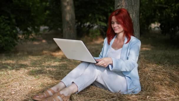 freelancing, joven empresaria trabajando remotamente y tecleando en un teclado de computadora en un parque sentado cerca de un árbol - Metraje, vídeo