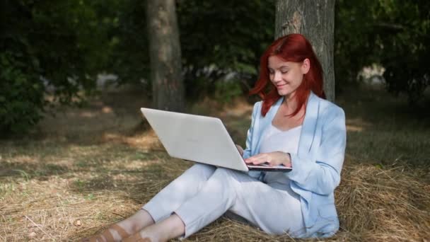 outdoor work, beautiful freelancer girl works at computer sitting in clearing near tree in park - Footage, Video