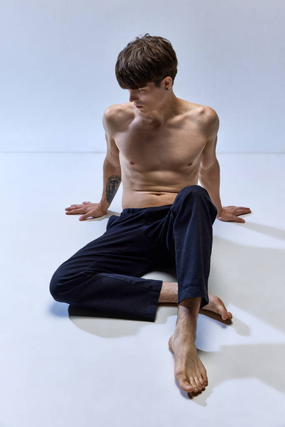 Full-length portrait of young half-naked man posing on floor isolated over grey studio background. Male muscular build body. Concept of mens health, beauty, fashion, body and skin care, wellness. ad - Photo, Image