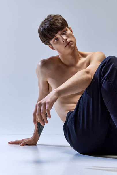 Porträt eines jungen attraktiven brünetten Mannes, der isoliert auf dem Boden vor dunkelgrauem Studiohintergrund sitzt. Männlicher muskulöser Körper. Konzept der Männergesundheit, Schönheit, Mode, Körper- und Hautpflege, Wellness - Foto, Bild