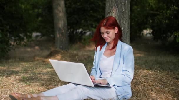 independiente, encantadora joven mujer que trabaja en un ordenador portátil al aire libre sentado en el claro en el parque entre los árboles - Imágenes, Vídeo