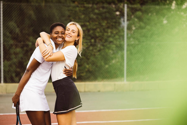 Retrato de dos jóvenes felices hermosas mujeres con ropa de tenis y raquetas en una cancha de tenis listo para jugar un juego. Dos amigos compartiendo una mañana de actividad deportiva jugando un partido de tenis. - Foto, Imagen