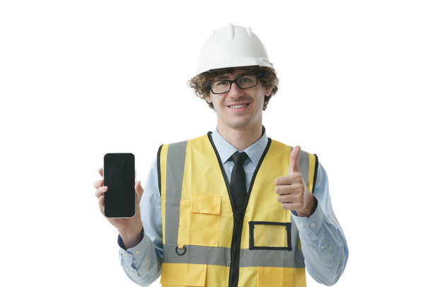 Ritratto di giovane operaio industriale, ingegnere con il cappello duro sorridente che mostra il telefono con il pollice in su, guardando felice e sicuro di sé, professione professionale. - Foto, immagini