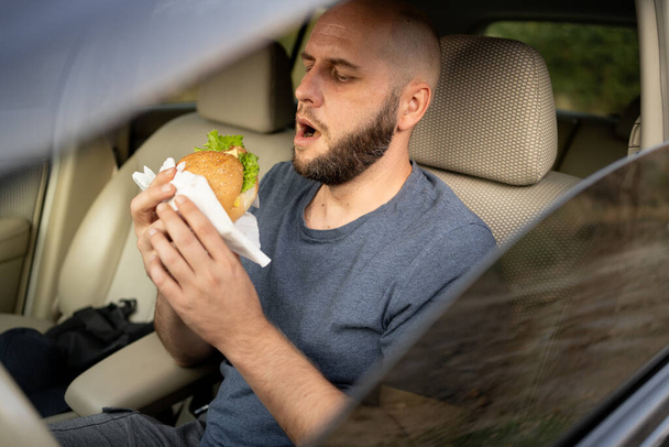Hombre comiendo hamburguesa en el coche. Tómate un descanso. Descansando en el viaje por carretera. Cena en automóvil. Asiento delantero. Concepto de comida. Copiar espacio - Foto, imagen