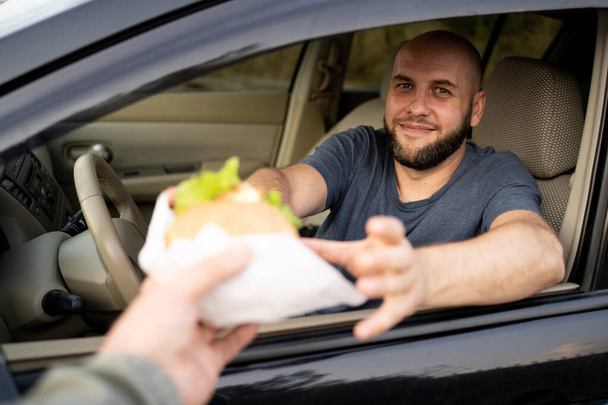 O correio de entrega de comida dá o hambúrguer ao homem no carro. Alimentos de segurança entregues. Um hambúrguer humano pela janela do carro. Espaço de cópia - Foto, Imagem