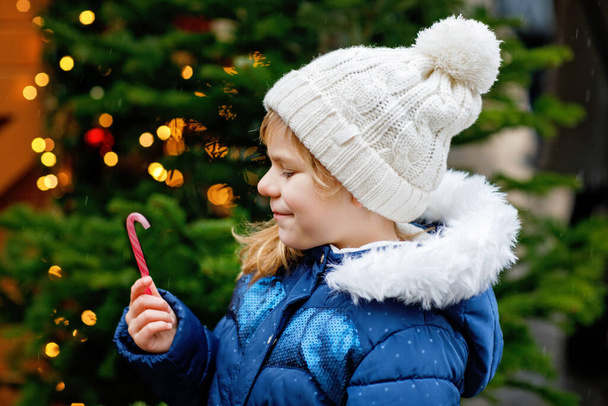 Kleine süße Vorschulmädchen mit Zuckerrohr vom Süßwarenstand auf dem Weihnachtsmarkt. Glückliches Kind auf dem traditionellen Familienmarkt in Deutschland. Vorschulkinder in bunter Winterkleidung bei Schneefall. - Foto, Bild