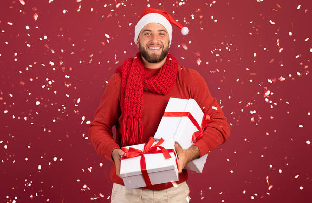 Счастливый миллениал-кавказец в шляпе Санты держит коробки с подарками, наслаждается праздниками со снегом, изолированным на фоне бургундской студии. Новогодняя вечеринка с конфетти - Фото, изображение