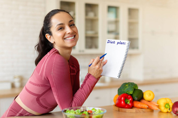 Hymyilevä sovi nainen poseeraa ruokavalio suunnitelma, kirjoittaminen valikko ja terve laihtuminen reseptit kannettavassa, seisoo moderni keittiö sisätiloissa, katsot kameraa. Laihdutus ravitsemus käsite - Valokuva, kuva
