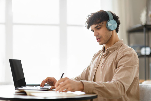 Vista lateral do jovem empresário árabe homem no laptop Notas de escrita vestindo fones de ouvido sem fio, trabalhando on-line e aprendendo na mesa de escritório interior. Conceito moderno de tecnologia e pessoas - Foto, Imagem
