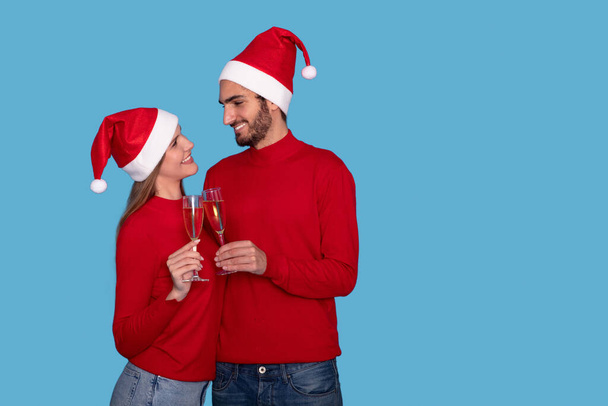Gelukkig romantisch paar in Santa hoeden klinkende champagne glazen en kijken naar elkaar, liefhebbende man en vrouw vieren wintervakantie samen, staande geïsoleerd op blauwe achtergrond, kopiëren ruimte - Foto, afbeelding