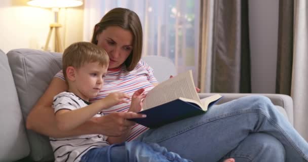 Moeder en zoon lezen een boek zittend op een gezellige bank in de woonkamer, familietijd. Hoge kwaliteit 4k beeldmateriaal - Video