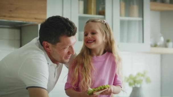 Uma menina sorridente alegre com uma sanduíche nas mãos fica na cozinha ao lado de seu pai. O pai com humor de brincadeira conta histórias engraçadas ou anedotas à filha. O conceito de amizade, feliz - Filmagem, Vídeo