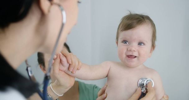 Kinderarzt legt Stethoskop auf Rücken des hübschen kleinen Kindes, um Herzschlag und Lunge zu hören. Mutter hält ihr süßes Baby beim Arztbesuch an den Händen. Arzt arbeitet in modernem Krankenhaus. Nahaufnahme. - Foto, Bild