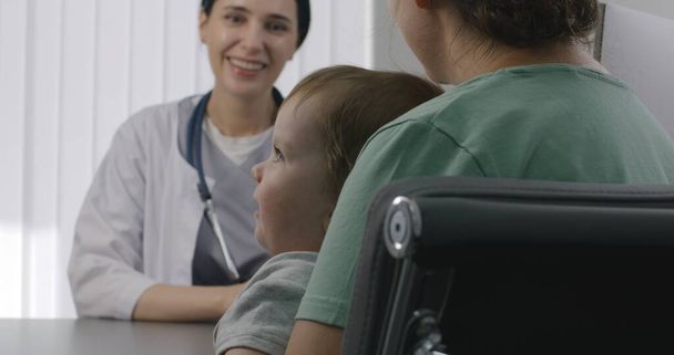 Kinderärztin berät Patientin mit süßem Kleinkind auf Termin im Krankenhaus. Frau sitzt auf Stuhl bei Arztuntersuchung. Freundlicher Arzt arbeitet in modernem medizinischen Zentrum. Aus nächster Nähe. Rückseite. - Foto, Bild