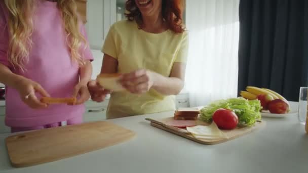 Hymyilevä äiti ja tytär keittiössä taitto voileipiä uhkapeli tuulella. Ruoanlaittoprosessi leikkisällä tavalla. Vanhempien ja lasten välinen ystävyys. Käsite onnellinen lapsuus ja - Materiaali, video