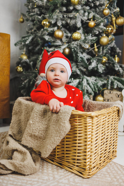 Ένα αγοράκι ενός έτους κοντά στο χριστουγεννιάτικο δέντρο. μια ιδέα για μια παιδική φωτογραφία για το νέο έτος. διακόσμηση στούντιο το νέο έτος. Χριστουγεννιάτικα δώρα, εορταστική ενδυμασία κόκκινων παιδιών - Φωτογραφία, εικόνα