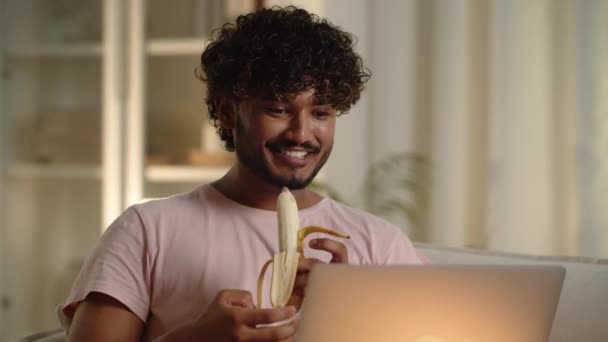 Un joven árabe con el pelo rizado desayunando viendo películas. Hombre feliz comiendo plátano al ver la conferencia en la computadora. Masculino estudiante almuerzo relajante mientras que a distancia estudiar buscando portátil en - Metraje, vídeo
