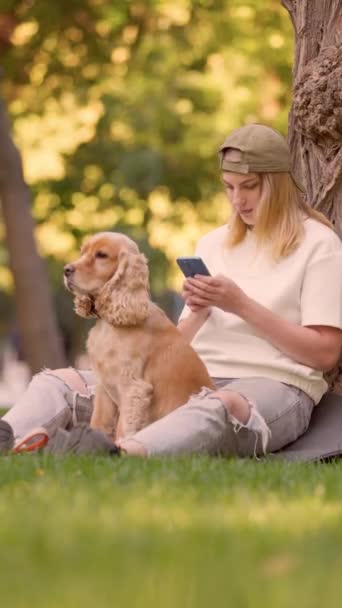 Νεαρή κοπέλα κάθεται στο γκαζόν στο πάρκο της πόλης με αγγλικά κόκερ σκυλί σπανιέλ και χρησιμοποιεί το τηλέφωνο - Πλάνα, βίντεο