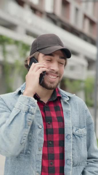 Młody człowiek w radosnym nastroju rozmawia przez telefon i uśmiecha się podczas spaceru ulicą City - Materiał filmowy, wideo