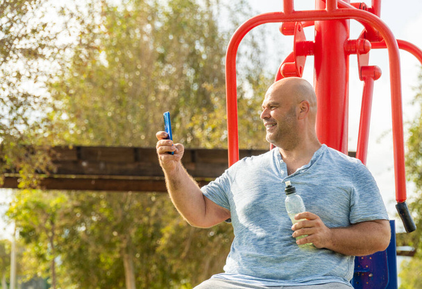 Egy férfi használja a mobilját, amint a gépen ül a bioszalibilis parkban, edzés után. - Fotó, kép