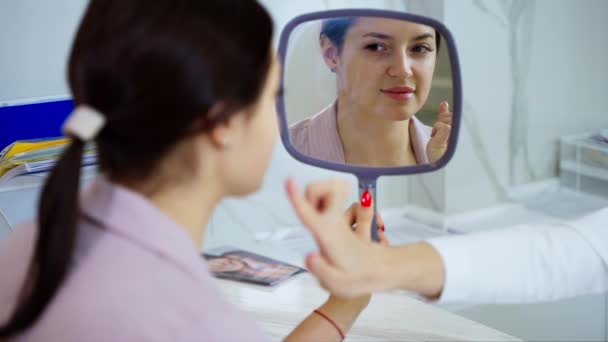 kosmetologinen klinikka, aikuinen nainen tapaamisessa estetiikan ja plastiikkakirurgin kanssa, kuuleminen kosmetologin kanssa, kasvojen ihon katselu peilissä - Materiaali, video