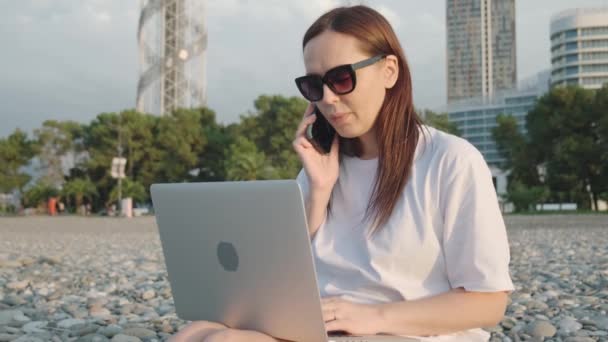 Digitální kočovnice veselá žena pracující s laptopem a mluvící na mobilním telefonu na pláži u moře. Žena na volné noze práci on-line pomocí smartphonu a počítače z dálky. Vzdálená práce na - Záběry, video