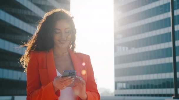 Щаслива молода змішана жінка, яка друкує повідомлення в чаті на смартфоні з золотим заходом сонця на фоні. Усміхнена бізнес-леді використовує інформацію про пошук мобільного телефону на бездротовому Інтернеті, онлайн-покупки 4K - Кадри, відео