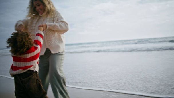 Veselá matka točí synem na břehu oceánu. Šťastná rodina hrající oceánskou pláž odpočívající na podzimní dovolenou. Krásná usměvavá chůva víření baví s malým kudrnatý chlapec. Radostný společný čas - Fotografie, Obrázek