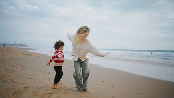 Η οικογένεια παίζει τον πιλότο του αεροπλάνου στην ακτή του ωκεανού. Χαρούμενη μητέρα γιος τρέχει παραλία διασκεδάζοντας μαζί το Σαββατοκύριακο. Αξιολάτρευτο σγουρό αγόρι που αγκαλιάζει τη νεαρή μαμά. Παιχνιδιάρης γονιός απολαμβάνοντας το χρόνο με το παιδί. - Φωτογραφία, εικόνα