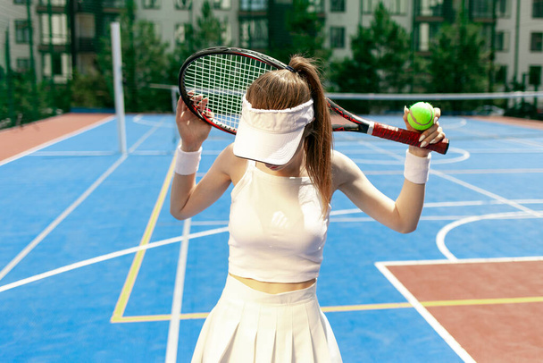young girl tennis player in white uniform holding racket on tennis court, female athlete playing tennis outdoors - Photo, Image