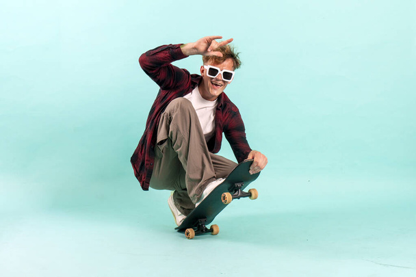 young cheerful guy rides skateboard on blue isolated background, hipster skater in sunglasses shows rock gesture - Photo, Image